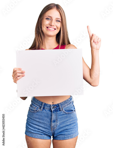 Young beautiful blonde woman holding blank empty banner surprised with an idea or question pointing finger with happy face, number one