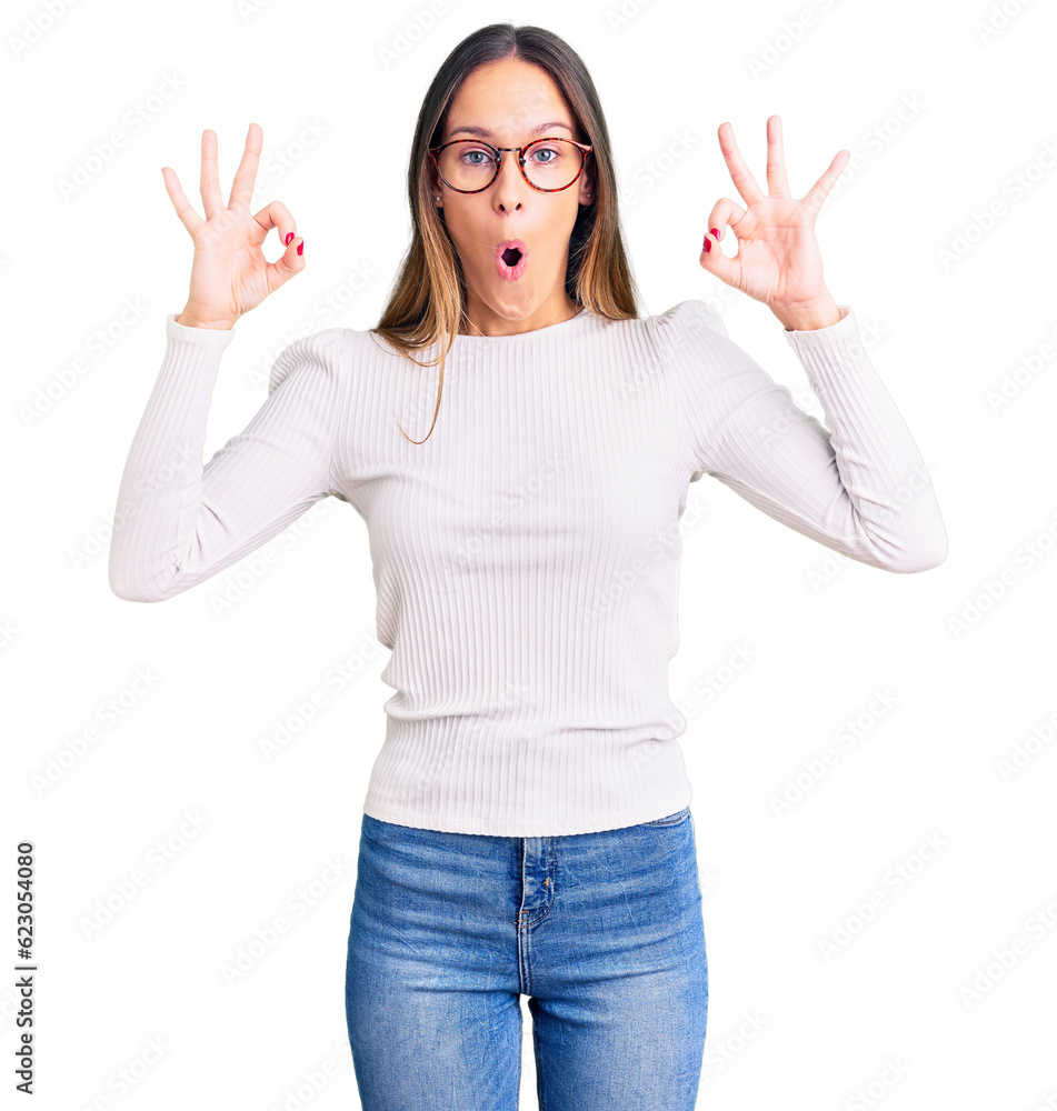 Beautiful brunette young woman wearing casual white sweater and glasses looking surprised and shocked doing ok approval symbol with fingers. crazy expression Beautiful brunette young woman wearing casual white sweater and glasses looking surprised and shocked doing ok approval symbol with fingers. crazy expression