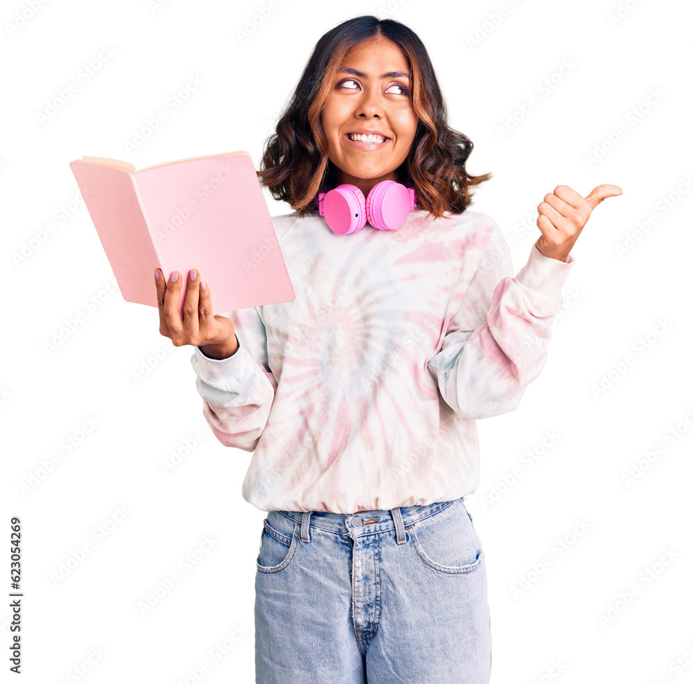 Young beautiful mixed race woman reading a book wearing headphones pointing thumb up to the side smiling happy with open mouth Young beautiful mixed race woman reading a book wearing headphones pointing thumb up to the side smiling happy with open mouth