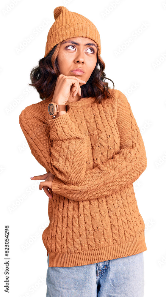 Young beautiful mixed race woman wearing wool sweater and winter hat with hand on chin thinking about question, pensive expression. smiling with thoughtful face. doubt concept. Young beautiful mixed race woman wearing wool sweater and winter hat with hand on chin thinking about question, pensive expression. smiling with thoughtful face. doubt concept.