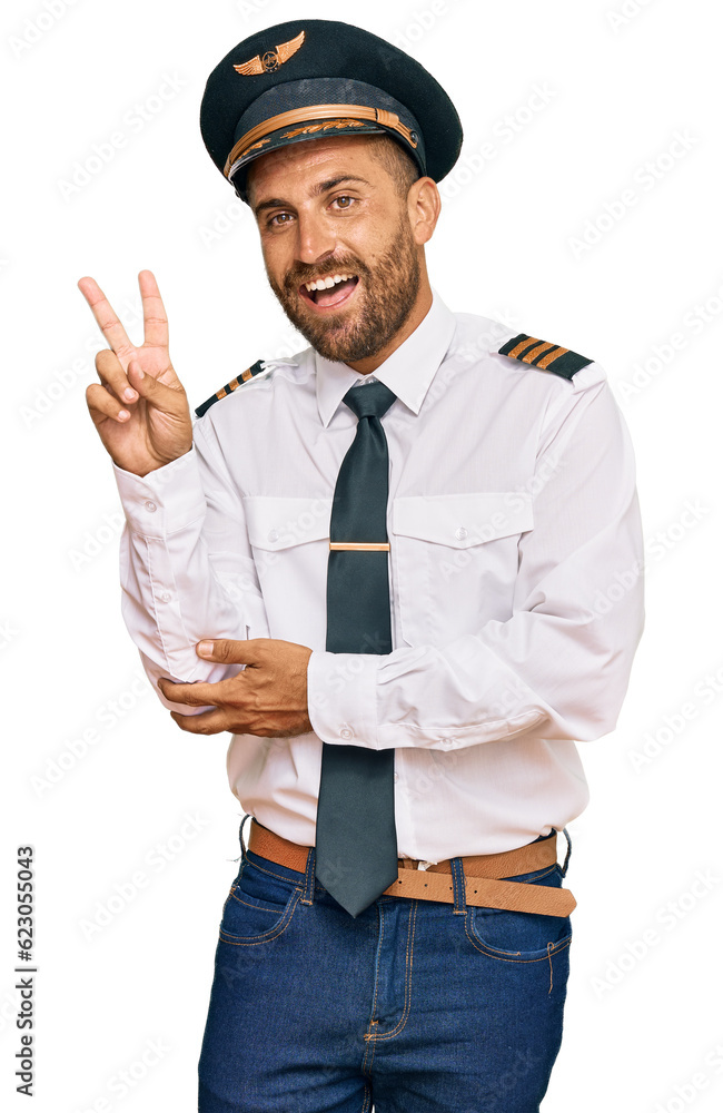 Handsome man with beard wearing airplane pilot uniform smiling with happy face winking at the camera doing victory sign. number two.