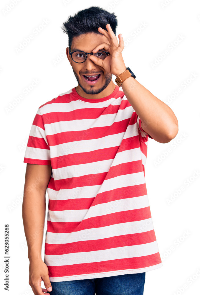 Handsome latin american young man wearing casual clothes and glasses doing ok gesture with hand smiling, eye looking through fingers with happy face. Handsome latin american young man wearing casual clothes and glasses doing ok gesture with hand smiling, eye looking through fingers with happy face.