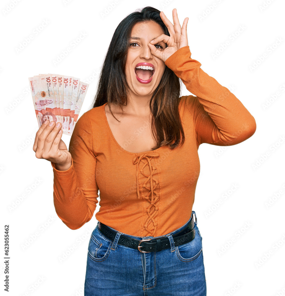 Beautiful hispanic woman holding 10 colombian pesos banknotes smiling happy doing ok sign with hand on eye looking through fingers Beautiful hispanic woman holding 10 colombian pesos banknotes smiling happy doing ok sign with hand on eye looking through fingers