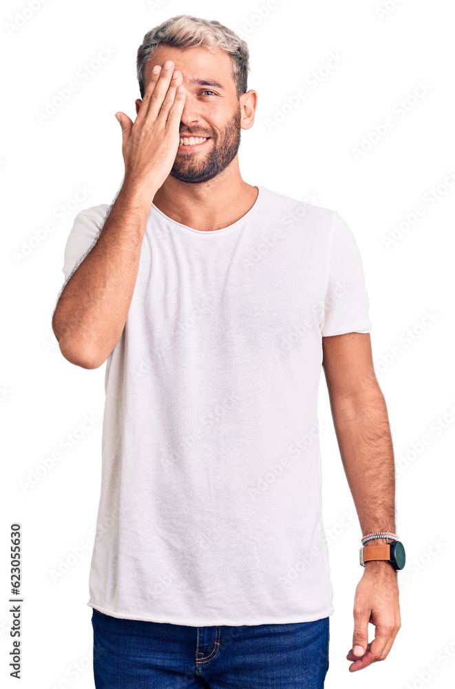 Young handsome blond man wearing casual t-shirt covering one eye with hand, confident smile on face and surprise emotion. Young handsome blond man wearing casual t-shirt covering one eye with hand, confident smile on face and surprise emotion.