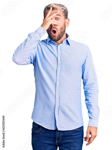 Young handsome blond man wearing elegant shirt peeking in shock covering face and eyes with hand, looking through fingers with embarrassed expression. © Krakenimages.com