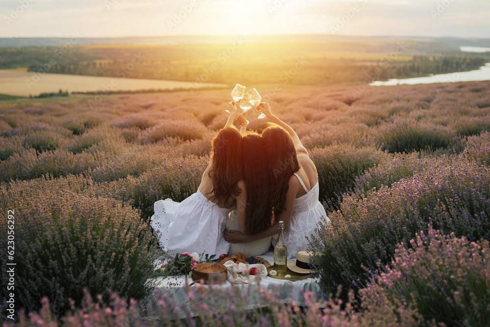 Girlfriends having picnic in the lavender field at sunset. Group of young women sitting on lavender field on summer day. Girlfriends drinking wine on outdoor party.