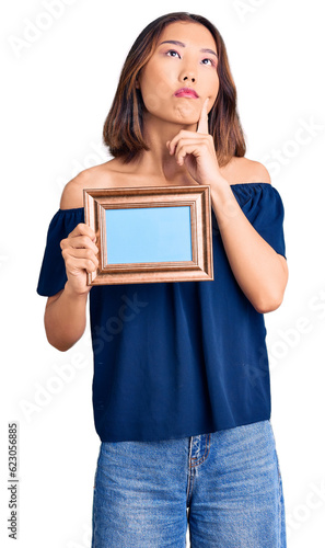 Young beautiful chinese girl holding empty frame serious face thinking about question with hand on chin, thoughtful about confusing idea