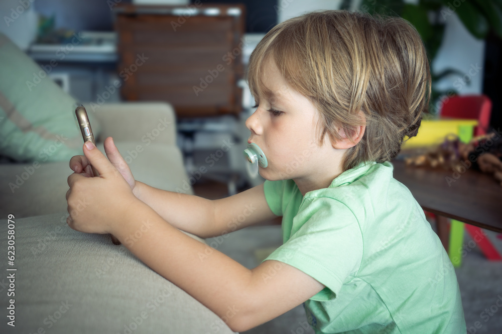 Close up cute little boy using smartphone, looking at screen, curious child holding phone in hands, sitting on couch at home alone, playing mobile device game, watching cartoons online. Close up cute little boy using smartphone, looking at screen, curious child holding phone in hands, sitting on couch at home alone, playing mobile device game, watching cartoons online.