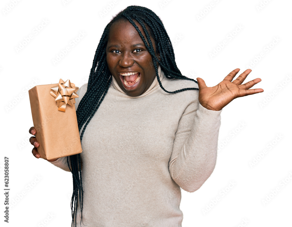 Young black woman with braids holding gift celebrating victory with happy smile and winner expression with raised hands Young black woman with braids holding gift celebrating victory with happy smile and winner expression with raised hands