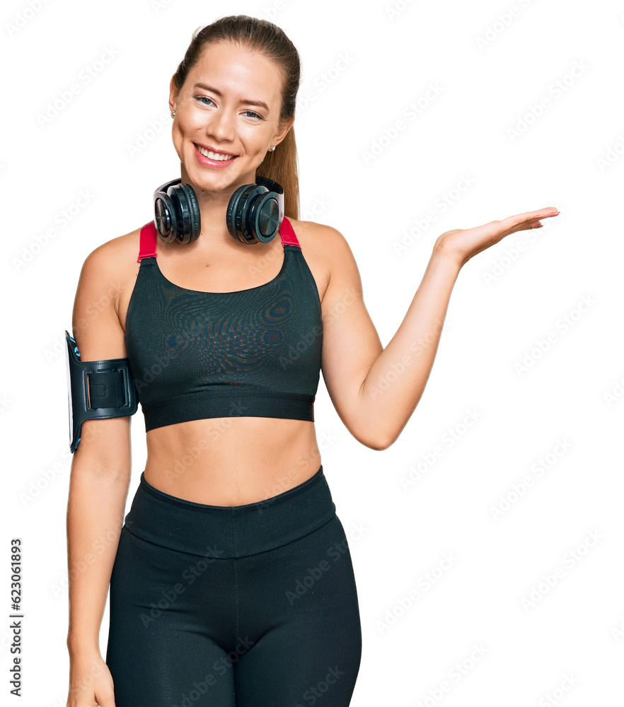 Beautiful blonde woman wearing gym clothes and using headphones smiling cheerful presenting and pointing with palm of hand looking at the camera. Beautiful blonde woman wearing gym clothes and using headphones smiling cheerful presenting and pointing with palm of hand looking at the camera.