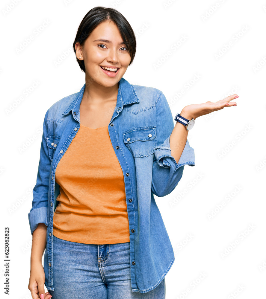 Young beautiful hispanic woman with short hair wearing casual denim jacket smiling cheerful presenting and pointing with palm of hand looking at the camera. Young beautiful hispanic woman with short hair wearing casual denim jacket smiling cheerful presenting and pointing with palm of hand looking at the camera.