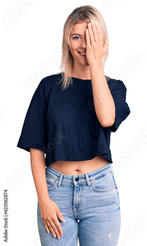 Young beautiful blonde woman wearing casual t-shirt covering one eye with hand, confident smile on face and surprise emotion.