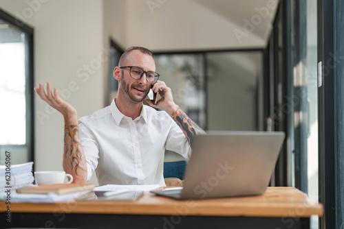Positive businessman talking on cellphone, sitting at workplace with laptop computer in modern office