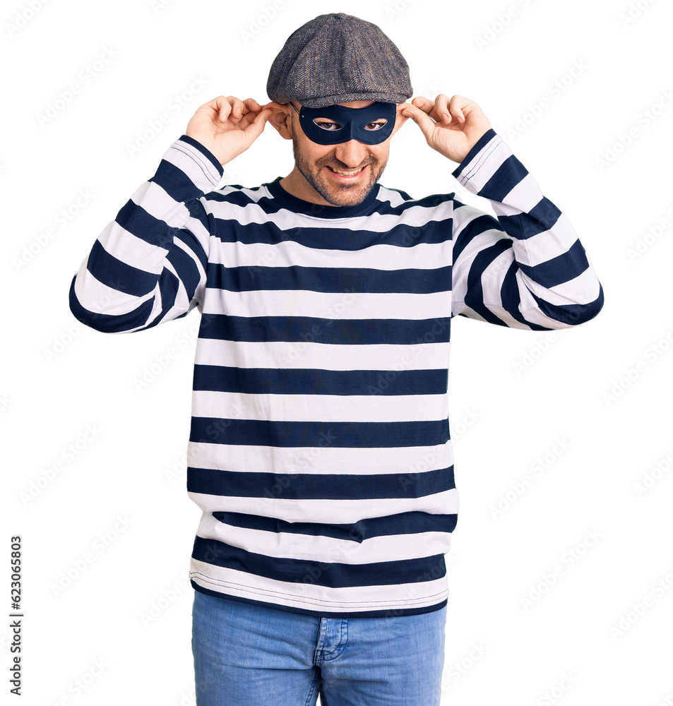 Young handsome man wearing burglar mask smiling pulling ears with fingers, funny gesture. audition problem Young handsome man wearing burglar mask smiling pulling ears with fingers, funny gesture. audition problem