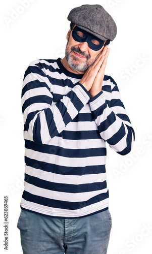 Middle age handsome man wearing burglar mask sleeping tired dreaming and posing with hands together while smiling with closed eyes. © Krakenimages.com