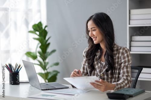 A beautiful Asian businesswoman sitting in her private office, she is checking company financial documents, she is a female executive of a startup company. Concept of financial management. © kamiphotos