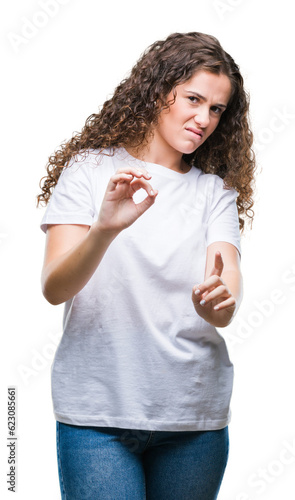 Beautiful brunette curly hair young girl wearing casual t-shirt over isolated background disgusted expression, displeased and fearful doing disgust face because aversion reaction. With hands raised