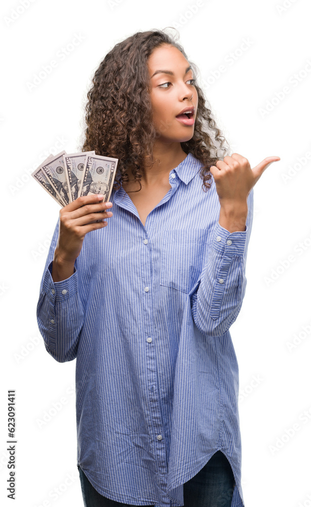 Young hispanic woman holding dollars pointing and showing with thumb up to the side with happy face smiling Young hispanic woman holding dollars pointing and showing with thumb up to the side with happy face smiling