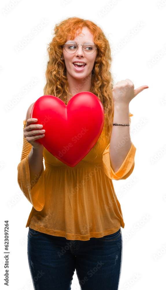 Young redhead woman in love holding red heart pointing and showing with thumb up to the side with happy face smiling Young redhead woman in love holding red heart pointing and showing with thumb up to the side with happy face smiling