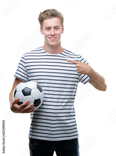 Young handsome blond man holding soccer ball with surprise face pointing finger to himself