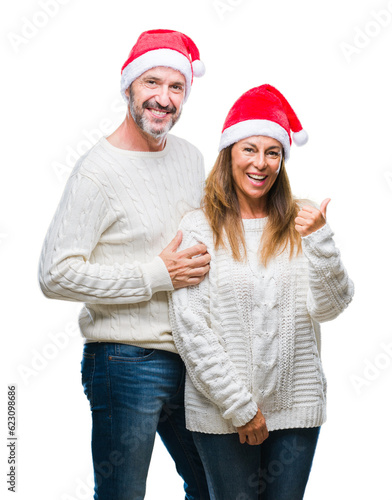 Middle age hispanic couple wearing christmas hat over isolated background smiling with happy face looking and pointing to the side with thumb up.