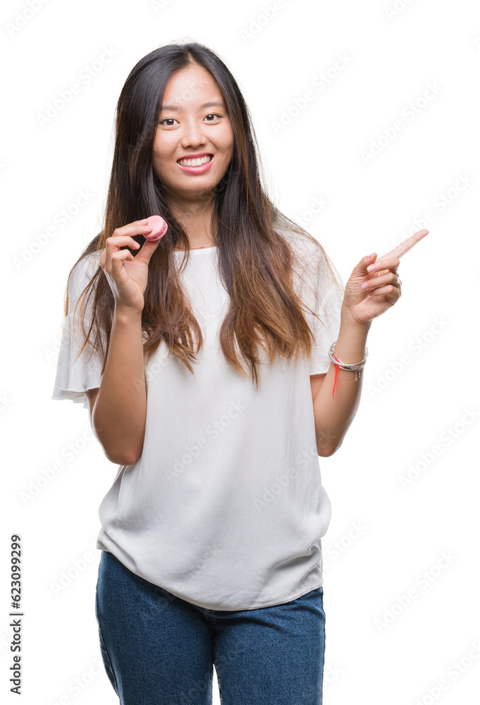 Young asian woman eating pink macaron sweet over isolated background very happy pointing with hand and finger to the side Young asian woman eating pink macaron sweet over isolated background very happy pointing with hand and finger to the side