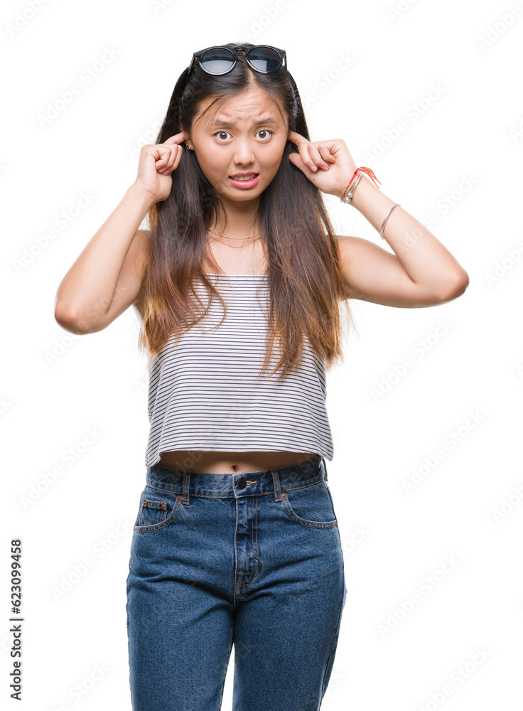 Young asian woman wearing sunglasses over isolated background covering ears with fingers with annoyed expression for the noise of loud music. Deaf concept. Young asian woman wearing sunglasses over isolated background covering ears with fingers with annoyed expression for the noise of loud music. Deaf concept.