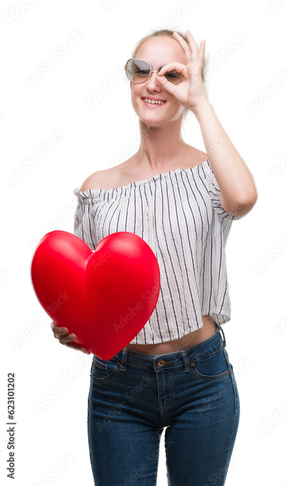 Blonde teenager woman holding red heart with happy face smiling doing ok sign with hand on eye looking through fingers Blonde teenager woman holding red heart with happy face smiling doing ok sign with hand on eye looking through fingers