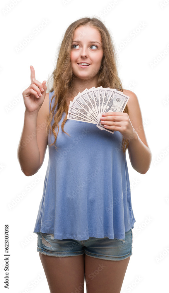 Obraz premium Young blonde woman holding dollars surprised with an idea or question pointing finger with happy face, number one