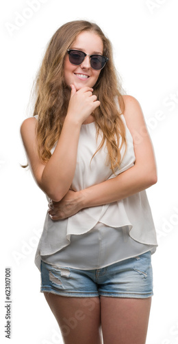 Young blonde woman wearing sunglasses looking confident at the camera with smile with crossed arms and hand raised on chin. Thinking positive.