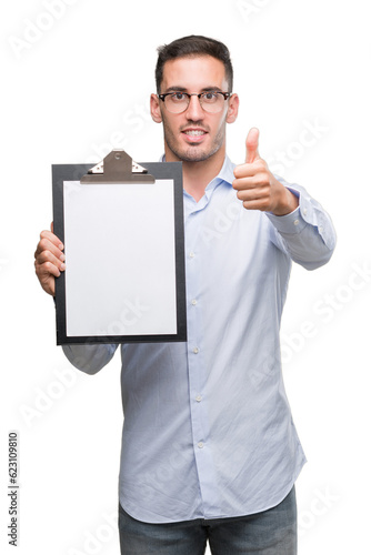 Handsome young business man showing a clipboard happy with big smile doing ok sign, thumb up with fingers, excellent sign
