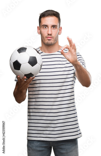 Handsome young man holding soccer football doing ok sign with fingers, excellent symbol