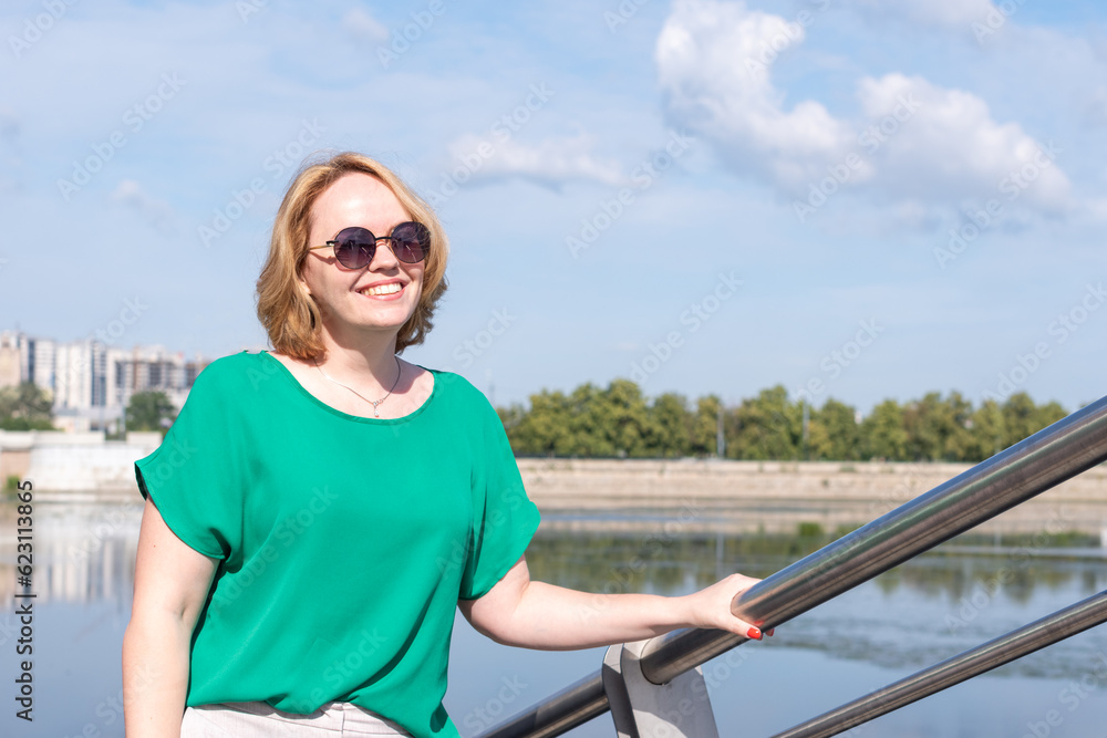 Portrait of a smiling happy middle-aged woman in sunglasses, green blouse standing on the river bank. A girl against the sky, a copy of space. A girl walks through the streets of the city.