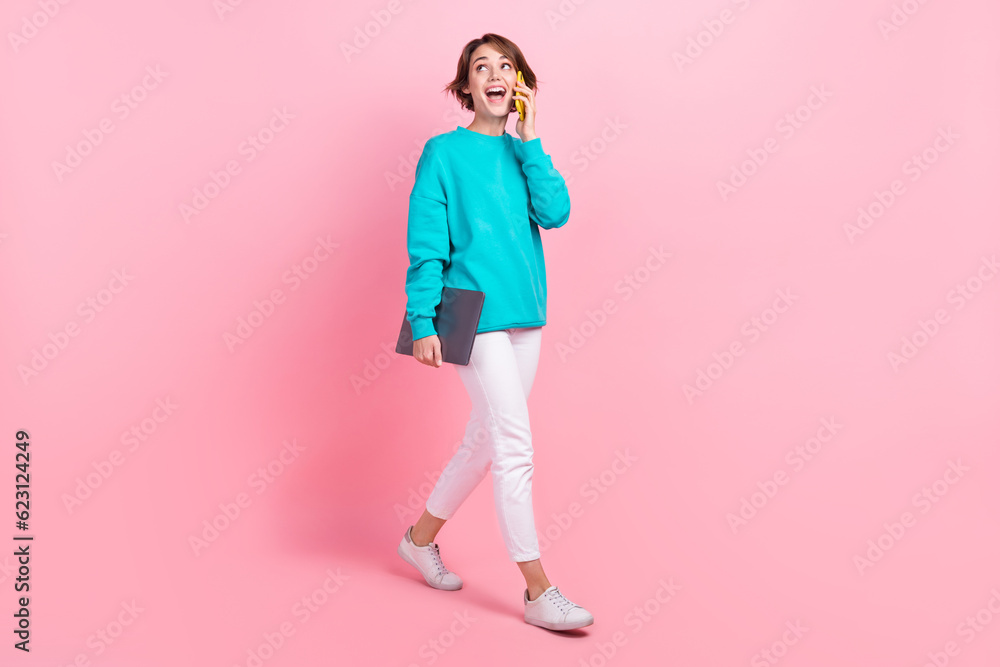 Full length photo of excited positive girl carry netbook speak phone look up empty space sale offer isolated on pink color background