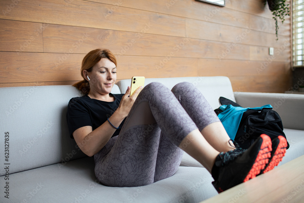 Obraz premium Mid adult woman using a smart phone at home while wearing her workout clothes