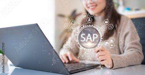 System Applications and Product (SAP). Program helps manage business to access information quickly accurately. Woman using laptop holding magnifying glass with SAP text inside. An icon on background. photo
