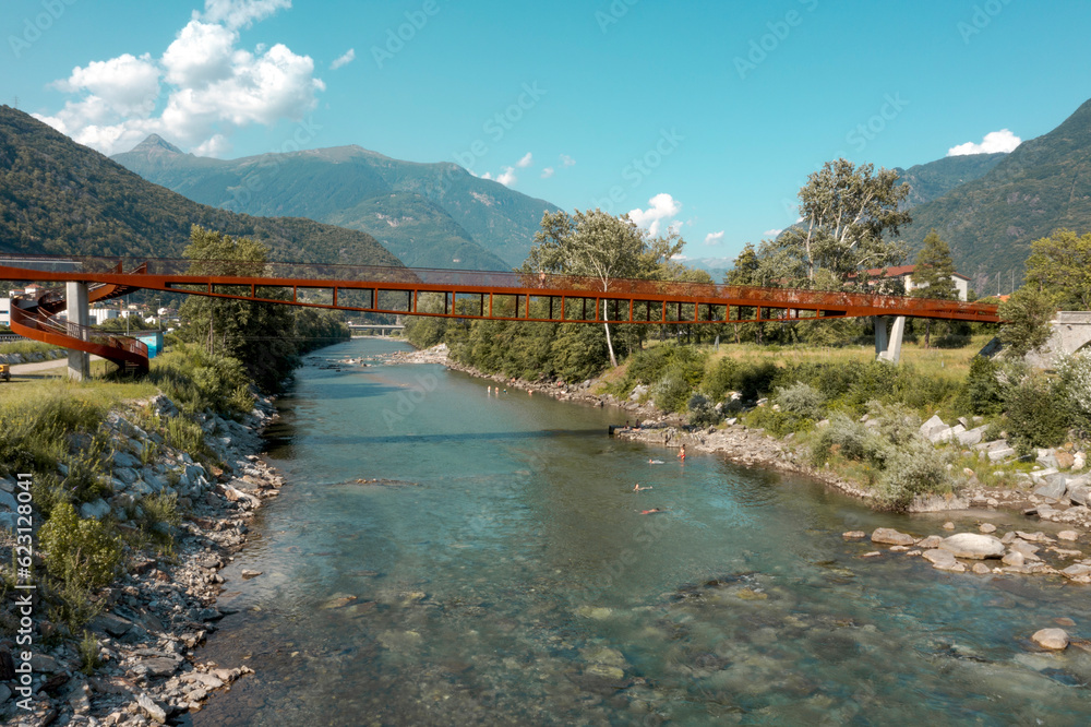 Fototapeta premium Modern rusted pedestrian and bicycle bridge with flowing Ticino river below. Beautiful panorama of the Alps of Italian Switzerland, taken from the sky