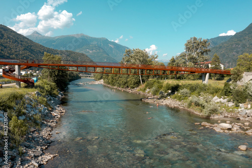 Modern rusted pedestrian and bicycle bridge with flowing Ticino river below. Beautiful panorama of the Alps of Italian Switzerland, taken from the sky