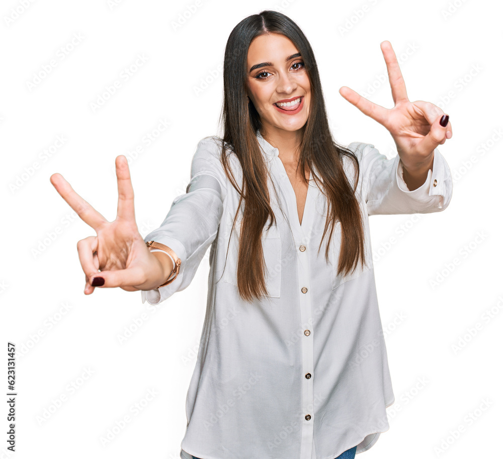 Young beautiful woman wearing casual white shirt smiling with tongue out showing fingers of both hands doing victory sign. number two. Young beautiful woman wearing casual white shirt smiling with tongue out showing fingers of both hands doing victory sign. number two.
