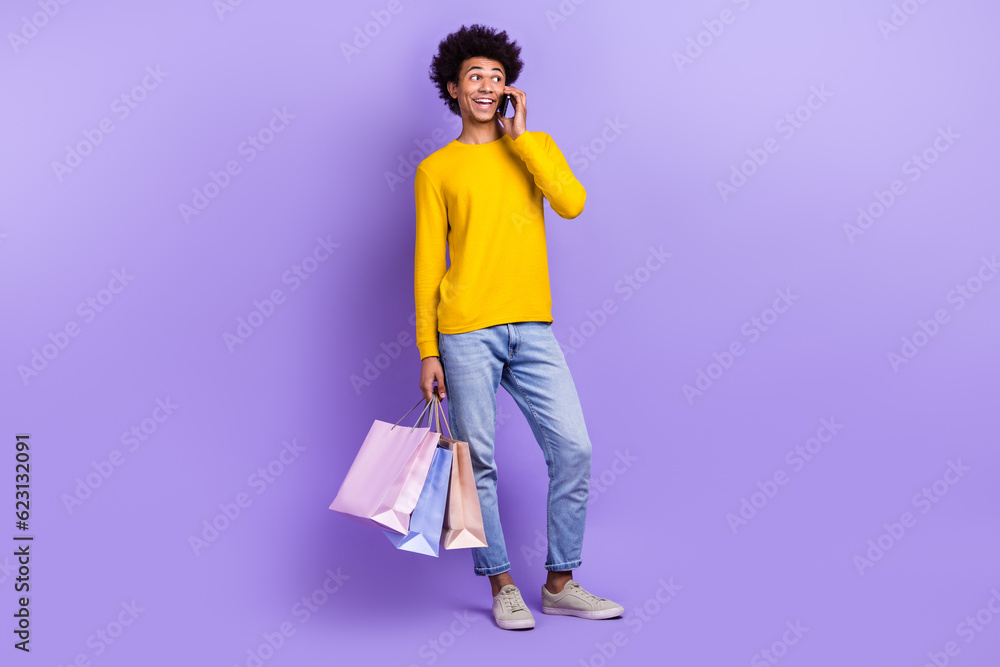 Full length photo of cheerful handsome man speak telephone hold store mall bags isolated on violet color background Full length photo of cheerful handsome man speak telephone hold store mall bags isolated on violet color background