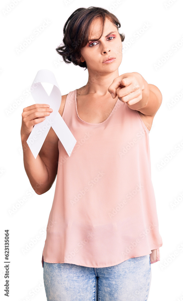 Beautiful young woman with short hair holding cancer awareness white ribbon pointing with finger to the camera and to you, confident gesture looking serious Beautiful young woman with short hair holding cancer awareness white ribbon pointing with finger to the camera and to you, confident gesture looking serious