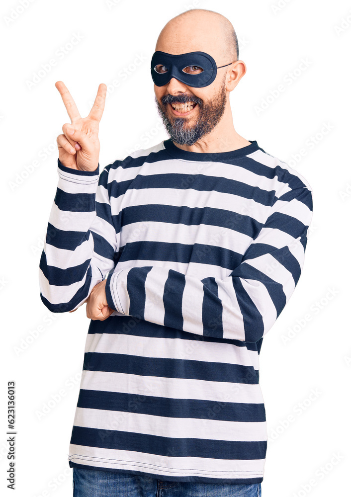Young handsome man wearing burglar mask smiling with happy face winking at the camera doing victory sign with fingers. number two. Young handsome man wearing burglar mask smiling with happy face winking at the camera doing victory sign with fingers. number two.