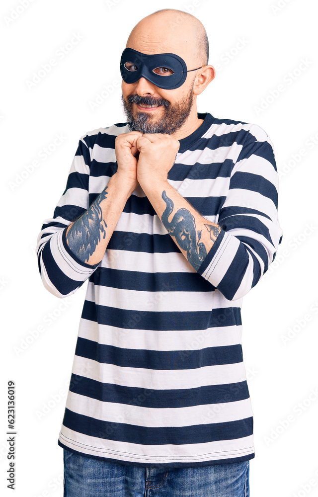 Young handsome man wearing burglar mask laughing nervous and excited with hands on chin looking to the side Young handsome man wearing burglar mask laughing nervous and excited with hands on chin looking to the side