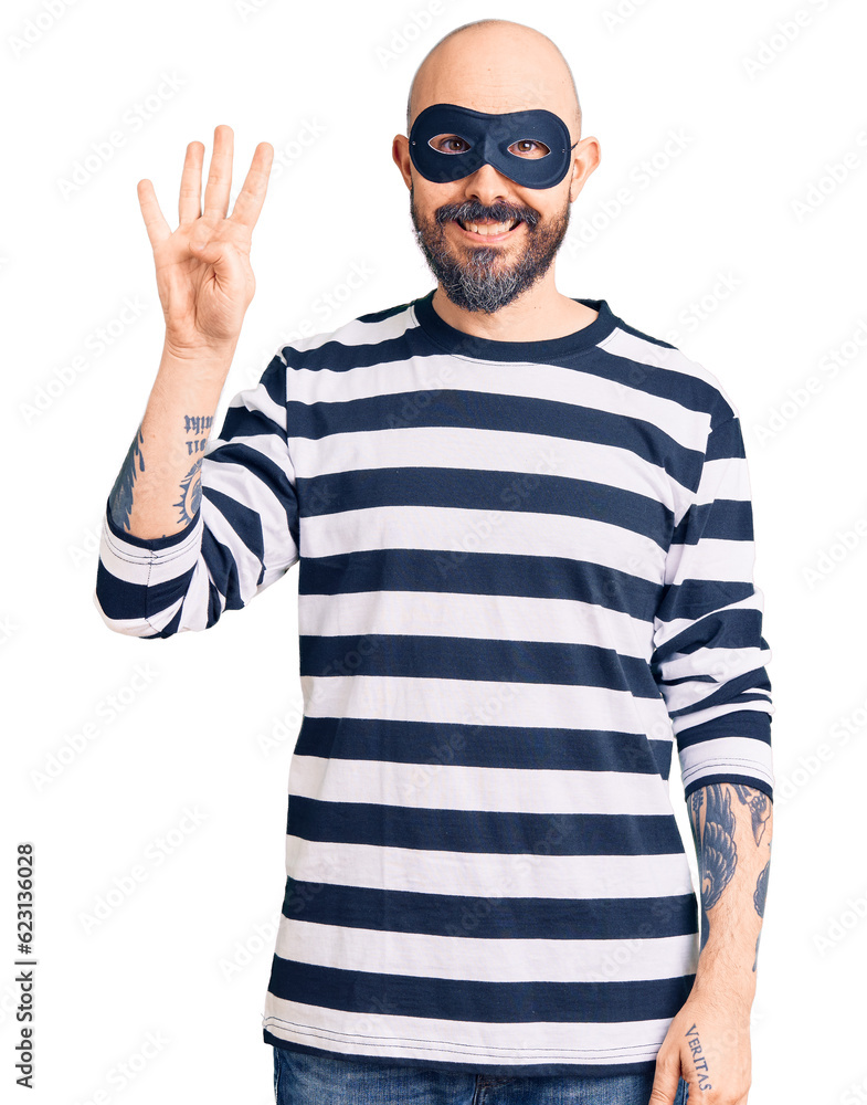Young handsome man wearing burglar mask showing and pointing up with fingers number four while smiling confident and happy. Young handsome man wearing burglar mask showing and pointing up with fingers number four while smiling confident and happy.