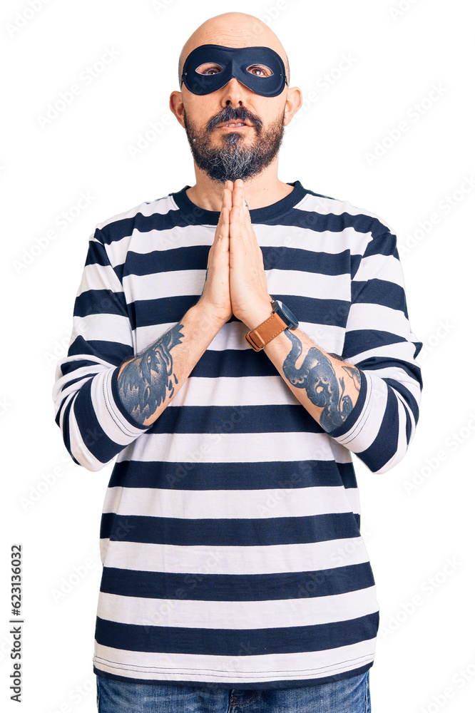 Young handsome man wearing burglar mask begging and praying with hands together with hope expression on face very emotional and worried. begging. Young handsome man wearing burglar mask begging and praying with hands together with hope expression on face very emotional and worried. begging.