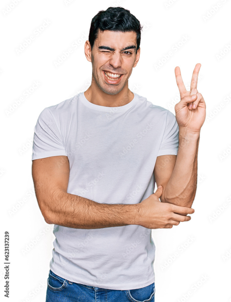 Naklejka premium Handsome hispanic man wearing casual white t shirt smiling with happy face winking at the camera doing victory sign. number two.
