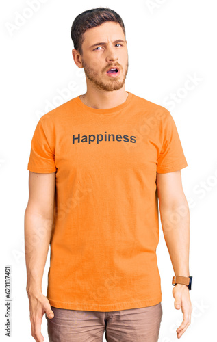 Handsome young man with bear wearing tshirt with happiness word message in shock face, looking skeptical and sarcastic, surprised with open mouth