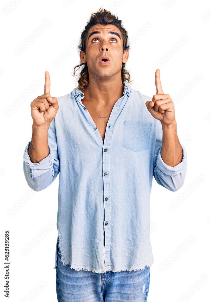 Young hispanic man wearing summer style amazed and surprised looking up and pointing with fingers and raised arms. Young hispanic man wearing summer style amazed and surprised looking up and pointing with fingers and raised arms.