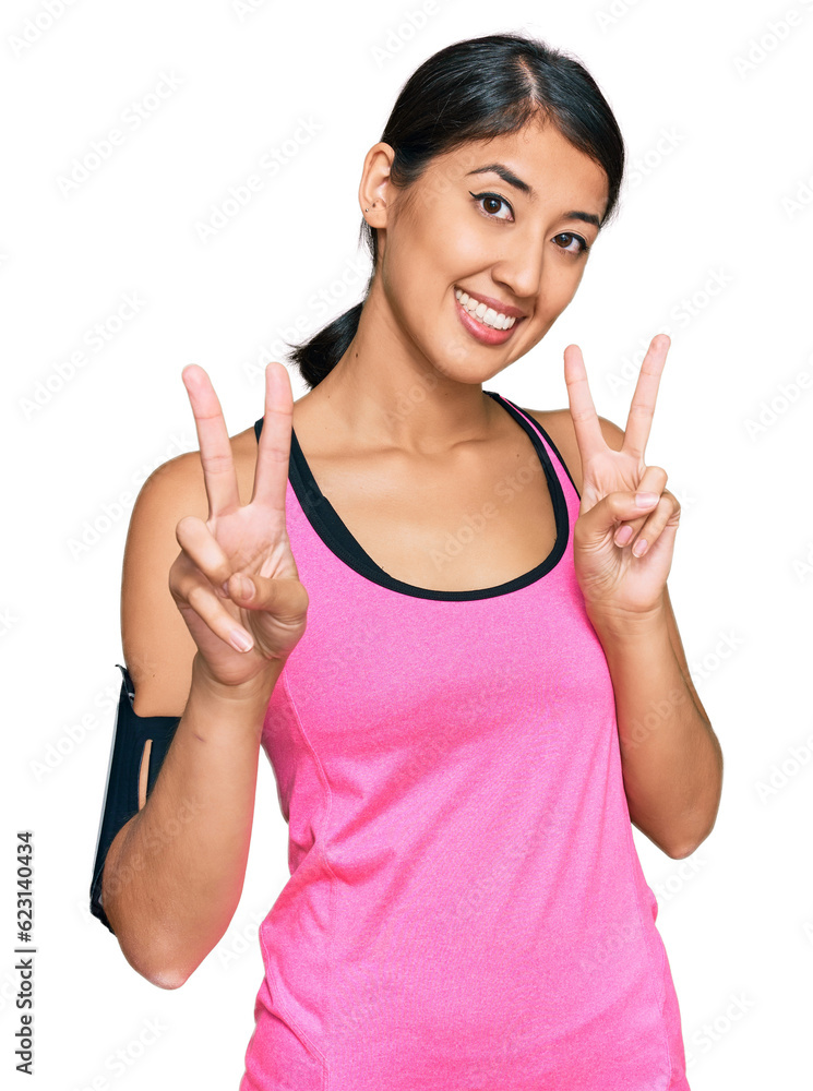 Beautiful asian young sport woman wearing sportswear and arm band smiling looking to the camera showing fingers doing victory sign. number two. Beautiful asian young sport woman wearing sportswear and arm band smiling looking to the camera showing fingers doing victory sign. number two.