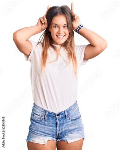Beautiful caucasian woman wearing casual white tshirt posing funny and crazy with fingers on head as bunny ears, smiling cheerful © Krakenimages.com